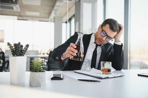 Vermoeidheid jonge zakenman die op de werkplek zit en een whiskyfles vasthoudt die alcohol faillissementsconcept drinkt