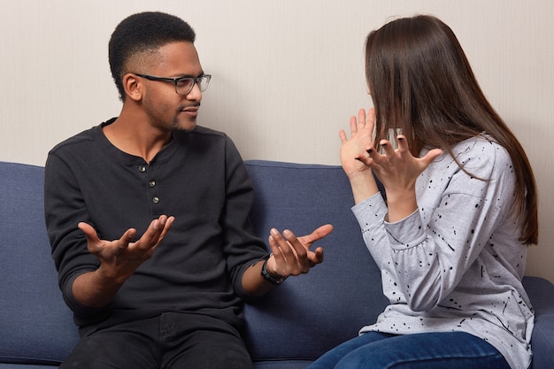 Verontwaardigd gebaar van vrouw en man boos, kijk naar elkaar, heb actieve ruzie, ben het ergens niet mee eens, voel je ontevreden, spreek hun mening uit, ga op een comfortabele bank zitten. Multi-etnisch gezinspaar betoogt