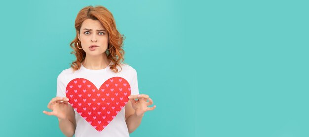 Foto verontwaardigd roodharig meisje houdt rood hart op blauwe achtergrond vrouw geïsoleerd gezicht portret banner met kopie