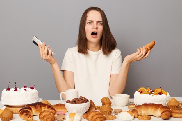 Verontwaardigde vrouw met bruin haar in wit T-shirt zittend aan tafel geïsoleerd over grijze achtergrond geschokt hoeveel calorieën ze at voor dag houdt zoet dessert vast en smartphone heeft geen dieet