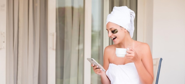 Verpakt in handdoek na het douchen vrouw met ooglapjes drinkt thee of koffie aan tafel en leest nieuws of berichten op het hotelterras meisje geniet van haar vakantie