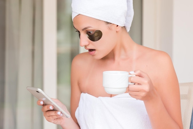 Verpakt in handdoek na het douchen vrouw met ooglapjes drinkt thee of koffie aan tafel en leest nieuws of berichten op het hotelterras Meisje geniet van haar vakantie