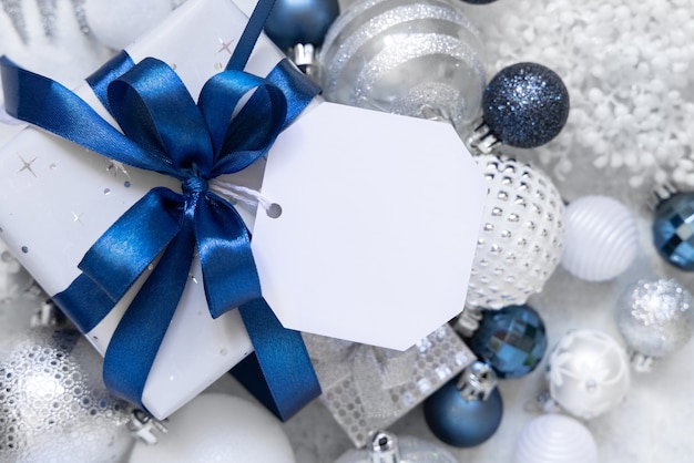 Verpakte geschenkdoos met een blauwe strik en papieren cadeaulabel op een witte tafel met witte en zilveren kerstversieringen rond het bovenaanzicht. Wintersamenstelling met blanco labelkaart, Mockup, kopieerruimte