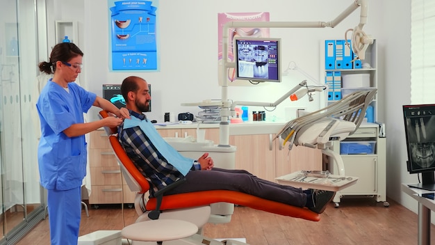 Foto verpleegkundige die tandheelkundige slabbetje aan de mens geeft vóór stomatologisch onderzoek, wachtend op stomatoloog-arts die de patiënt voorbereidt op een tandoperatie. verpleegkundige werkzaam in moderne orthodontische kliniek.