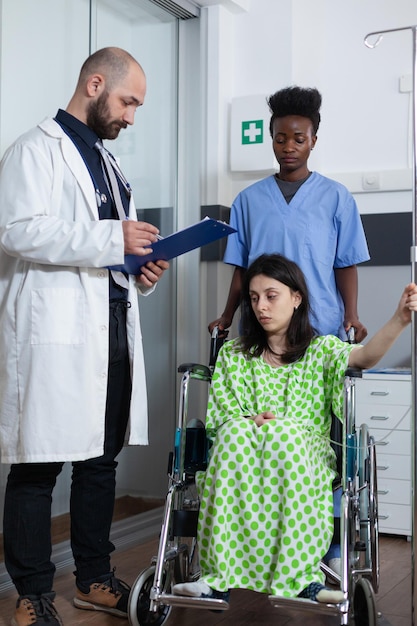 Verpleegkundige verplaatst patiënt zittend in rolstoel met iv-paal naar behandelkamer terwijl arts consult geeft en naar klembord kijkt met laboratoriumresultaten. Vrouw met ziekte die vóór de operatie wordt gecontroleerd.