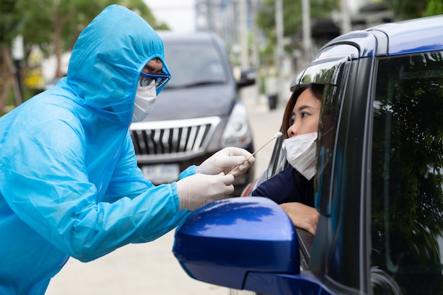 Verpleegster draagt een PBM-pak of medische hulpverleners in volledige beschermende kleding neemt een monster van de vrouwelijke bestuurder in de auto. Drive-thru-test voor Coronavirus COVID-19