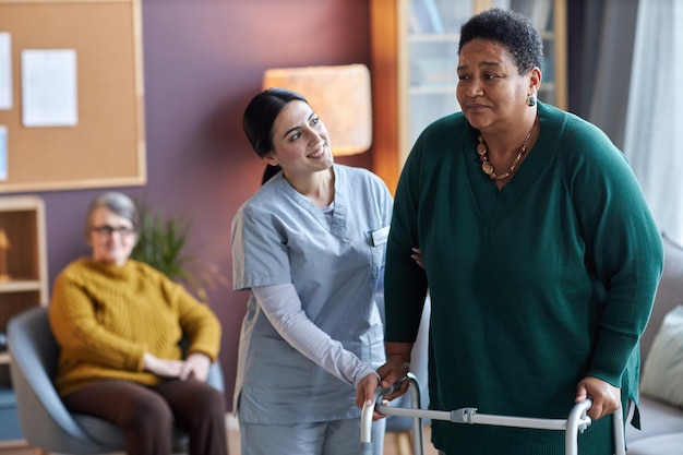 Verpleegster helpt zwarte oudere vrouw met behulp van mobiliteitswandelaar in bejaardentehuis