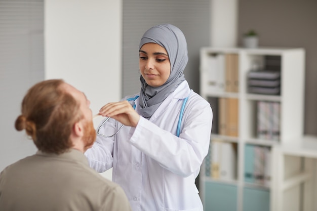 Verpleegster in hijab en in witte jas die de keel van de patiënt onderzoekt tijdens medisch onderzoek in het ziekenhuis