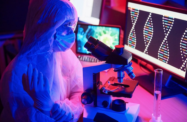 Verpleegster in masker en witte uniform zitten in neon verlichte laboratorium met computer, microscoop en medische apparatuur op zoek naar coronavirus vaccin