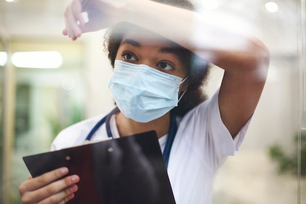 Verpleegster met masker die door het glas wegkijkt