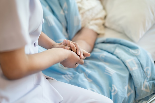 Verpleegster zittend op een ziekenhuisbed naast een oudere vrouw die handen helpt, zorg voor ouderen concept