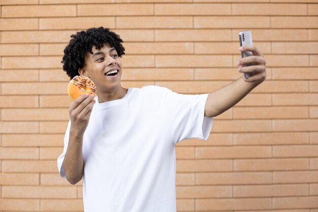 Verrast Afro-Amerikaanse man met krullend haar die selfies neemt met telefoon en een oranje donut vasthoudt