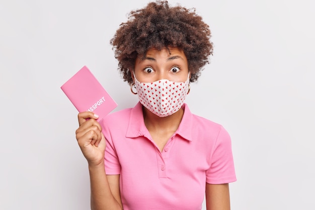 Verrast Afro-Amerikaanse vrouw met krullend haar draagt een beschermend hygiënemasker houdt paspoort vast dat gaat reizen tijdens de pandemie van het coronavirus en ontdekt enkele details over toekomstige vlucht geïsoleerd op een witte muur