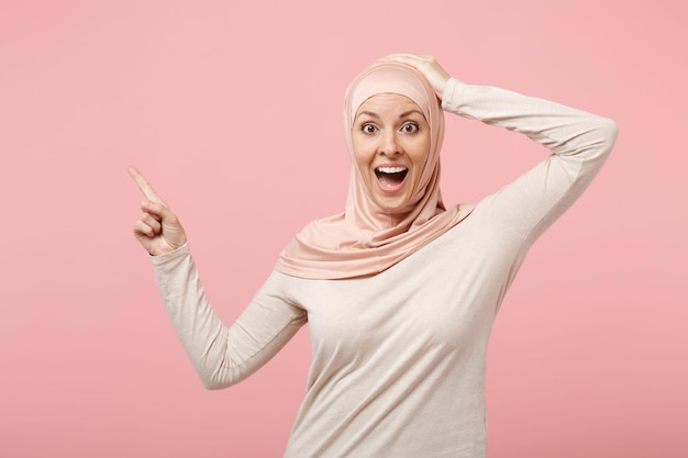 Verrast Arabische moslimvrouw in hijab lichte kleding poseren geïsoleerd op roze achtergrond. Mensen religieuze islam levensstijl concept. Bespotten kopie ruimte. Wijsvinger opzij wijzen, hand op het hoofd leggen.