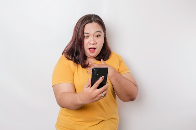 Verrast Aziatische grote vrouw met gele t-shirt wijzend op haar smartphone geïsoleerd door witte achtergrond