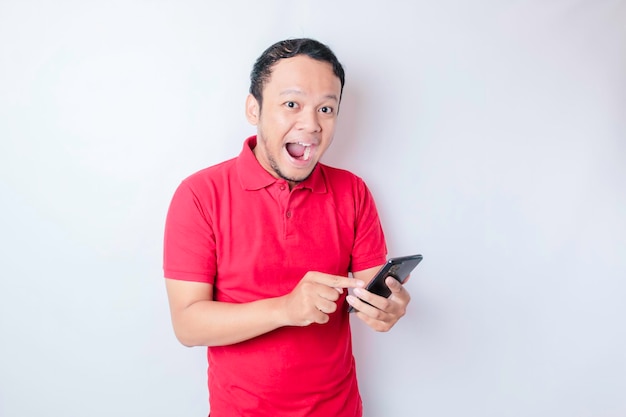 Verrast Aziatische man met rode t-shirt wijzend op zijn smartphone geïsoleerd door witte achtergrond
