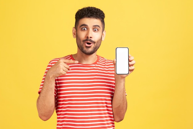 Verrast bebaarde man in gestreept t-shirt wijzende vinger naar smartphone met wit leeg display, geschokt door nieuwe telefoonfuncties. Indoor studio-opname geïsoleerd op gele achtergrond