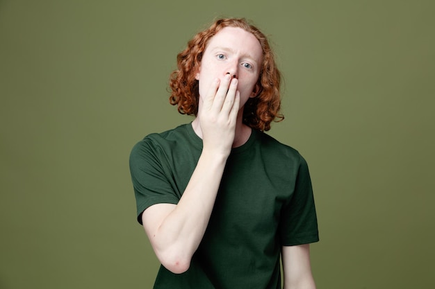 Verrast bedekt gezicht met hand jonge knappe jongen met groen t-shirt geïsoleerd op groene achtergrond