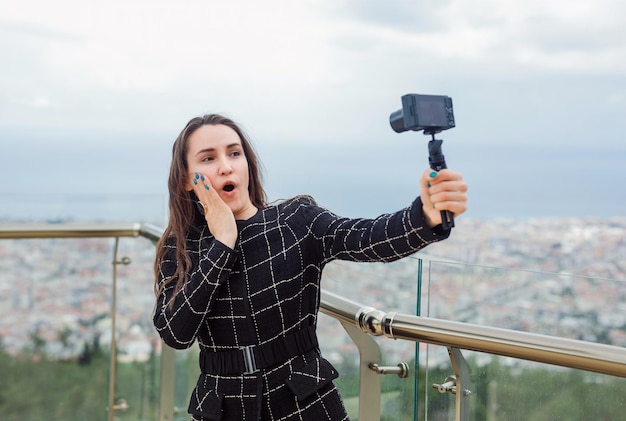 Verrast bloggermeisje neemt selfie door haar hand op de wang te houden tegen de achtergrond van het uitzicht op de stad