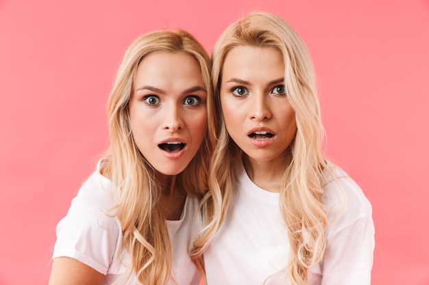 Verrast blonde tweeling gekleed in t-shirts kijken naar de voorkant over roze muur