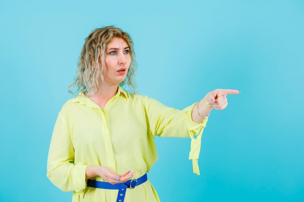 Foto verrast blonde vrouw wijst weg met wijsvinger op blauwe achtergrond