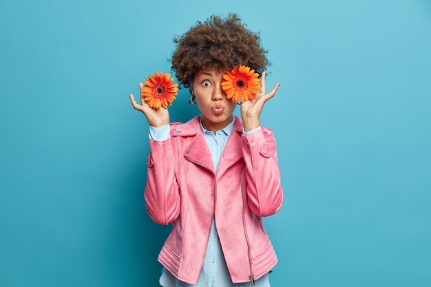 Verrast gekrulde jonge vrouw die perfect is als bloemen oranje gerberabloemen voor ogen houdt roze jas houdt lippen gevouwen geïsoleerd over blauwe muur