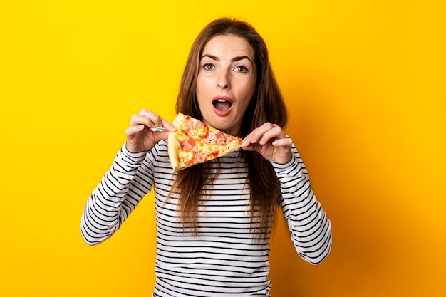 Verrast geschokte jonge vrouw kijkt met een plak hete verse pizza op een gele achtergrond