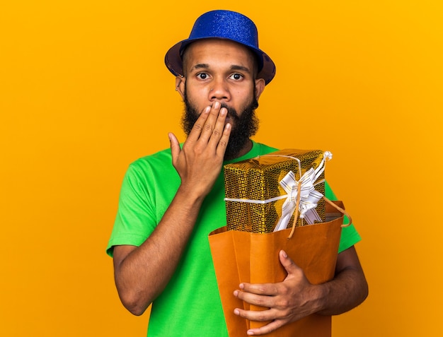 Verrast jonge Afro-Amerikaanse man met een feesthoed met een cadeauzakje bedekte mond met de hand geïsoleerd op een oranje muur