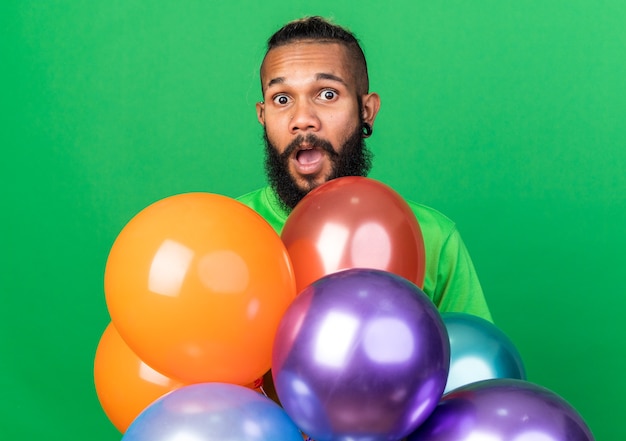 Verrast jonge Afro-Amerikaanse man met een groen t-shirt achter ballonnen geïsoleerd op een groene muur