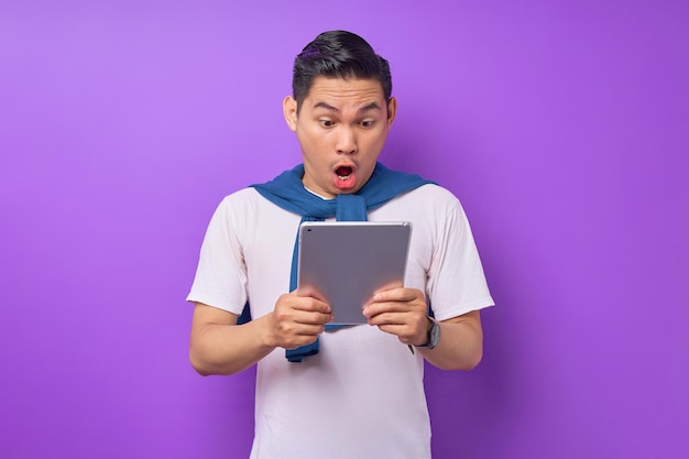 Verrast jonge Aziatische man in casual kleding met een digitale tablet en open mond geïsoleerd op paarse achtergrond