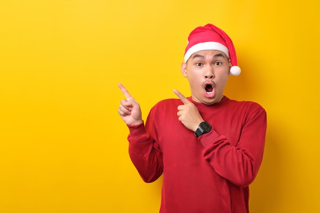 Verrast jonge Aziatische man in kerstmuts wijzende vingers opzij naar lege ruimte voor reclametekst op gele studio achtergrond viering kerstvakantie en nieuwjaarsconcept