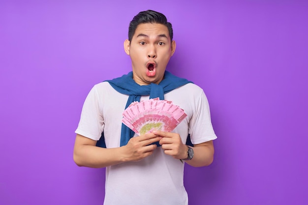Verrast jonge Aziatische man met witte t-shirt die geld bankbiljetten vasthoudt en camera geïsoleerd over paarse achtergrond bekijkt Winst en rijkdom concept