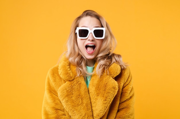 Verrast jonge blonde vrouw meisje in gele bontjas, donkere zonnebril poseren geïsoleerd op oranje achtergrond studio portret. Mensen emoties levensstijl concept. Bespotten kopie ruimte. Mond openhouden.