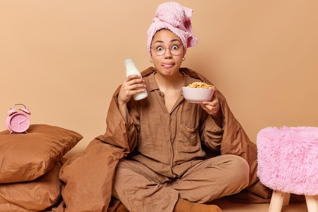 Verrast jonge Europese vrouw draagt pyjama houdt fles melk en kom cornflakes likt lippen voelt onder de indruk hoort schokkend nieuws geïsoleerd over beige achtergrond Ontbijt tijd concept