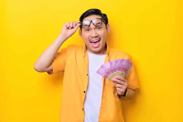Verrast jonge knappe Aziatische man die bankbiljetten vasthoudt en een bril afzet die op gele achtergrond is geïsoleerd