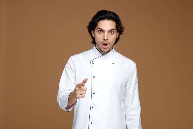 verrast jonge mannelijke chef-kok in uniform kijken en wijzend op camera geïsoleerd op bruine achtergrond