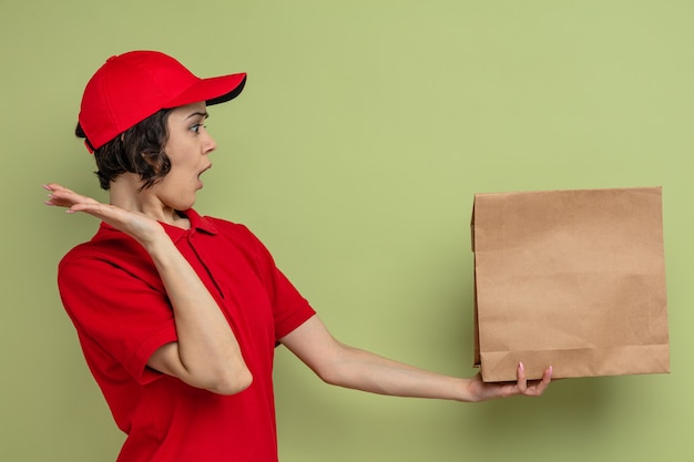 Verrast jonge, mooie bezorger die met opgeheven hand een papieren voedselzak vasthoudt en bekijkt