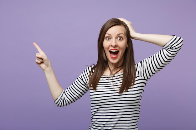 Verrast jonge vrouw in gestreepte kleding poseren geïsoleerd op violet paarse achtergrond. Mensen levensstijl concept. Bespotten kopie ruimte. De mond openhouden en de hand op het hoofd leggen en de wijsvinger opzij wijzen.