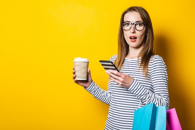 Verrast jonge vrouw met een telefoon, met boodschappentassen, met een kartonnen beker op een gele achtergrond