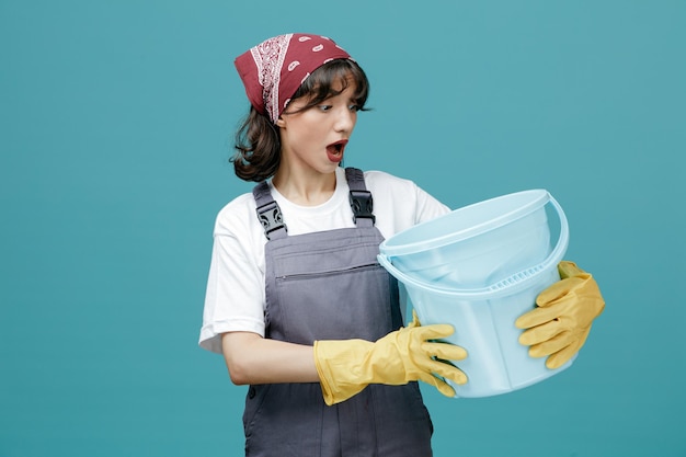 Verrast jonge vrouwelijke schoonmaker met uniforme bandana en rubberen handschoenen met emmer die ernaar kijkt geïsoleerd op blauwe achtergrond