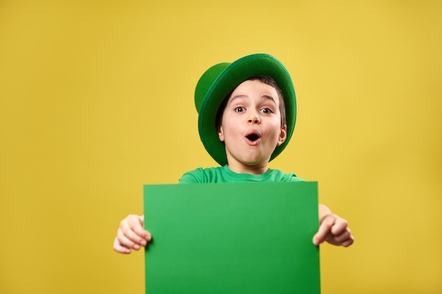 Verrast jongetje in groene Ierse kabouterhoed vormt met een groen vel papier in zijn handen