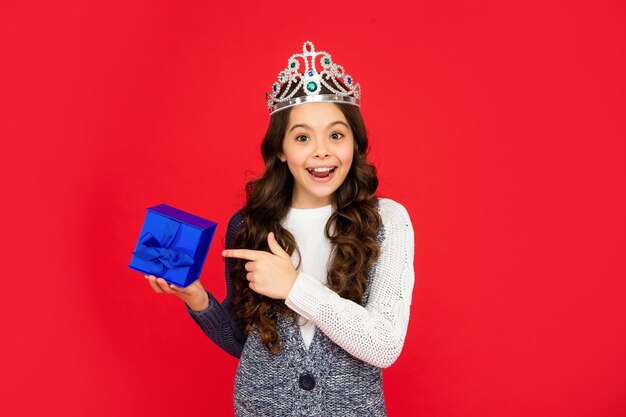 Verrast kind in koningin kroonprinses in tiara wijsvinger op doos kind met aanwezig tienermeisje draagt diadeem op rode achtergrond