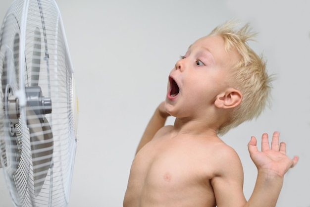 Verrast kleine blonde jongen met open mond en naakte torso staat in de buurt van een ventilator. Zomer concept