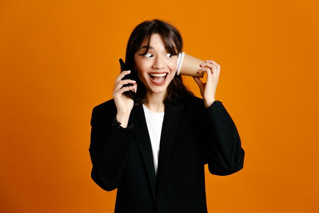 verrast koffiekopje op oor te zetten spreekt aan de telefoon jonge mooie vrouw draagt zwarte jas geïsoleerd op oranje achtergrond