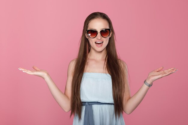 Verrast mode vrouw in zonnebril en blauwe jurk op roze achtergrond