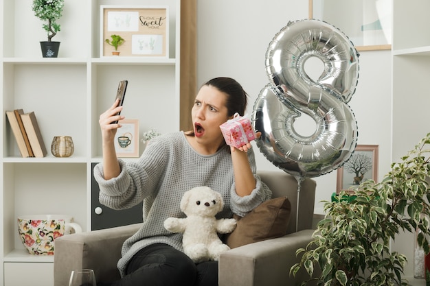 Verrast mooi meisje op een gelukkige vrouwendag die aanwezig is terwijl ze naar de telefoon in haar hand kijkt terwijl ze op een fauteuil in de woonkamer zit