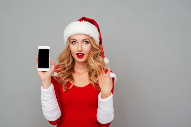 Verrast mooie jonge vrouw in nieuwjaarskostuum en hoed met lege scherm mobiele telefoon over grijs oppervlak