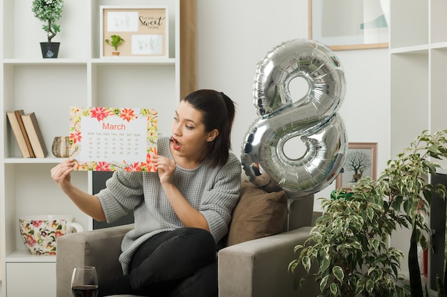 Verrast mooie vrouw op gelukkige vrouwendag houden en kijken naar kalender zittend op een fauteuil in de woonkamer