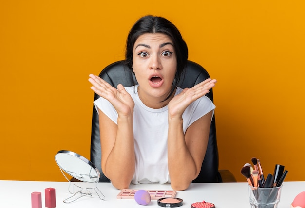 Verrast mooie vrouw zit aan tafel met make-up tools spreidende handen