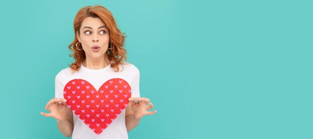 Foto verrast roodharige meisje houdt rood hart op blauwe achtergrond vrouw geïsoleerd gezicht portret banner met kopie ruimte